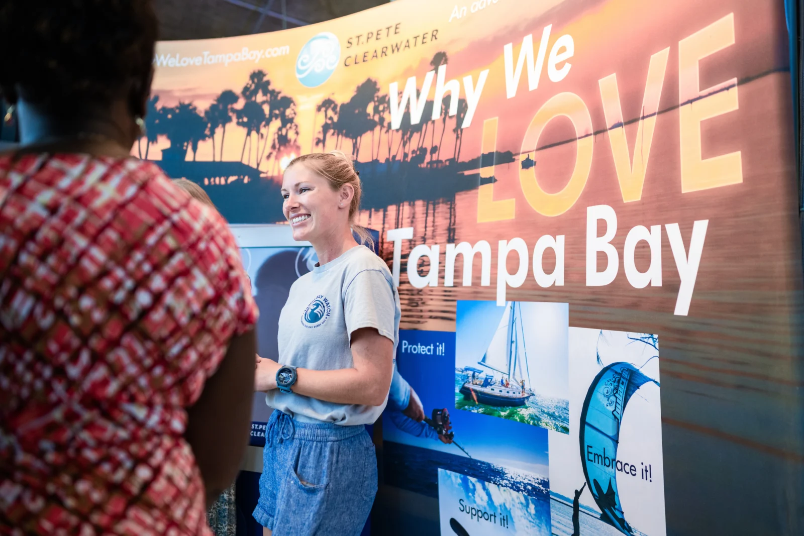 A Tampa Bay Watch staff member smiles while talking with visitors at a Why We Love Tampa Bay event booth.