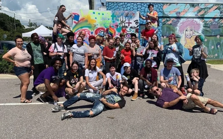 A large group of Diversity Arts participants poses in front of a colorful outdoor mural.