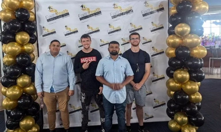 Four young men pose in front of a Faith Youth Services Inc. banner framed by black and gold balloon columns.
