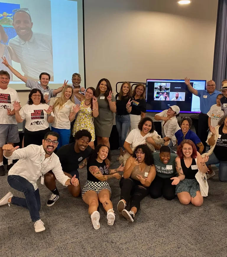 A cheering group of Mi Gente Mi Pueblo community members poses together in a meeting room, with more people joining on a video screen.
