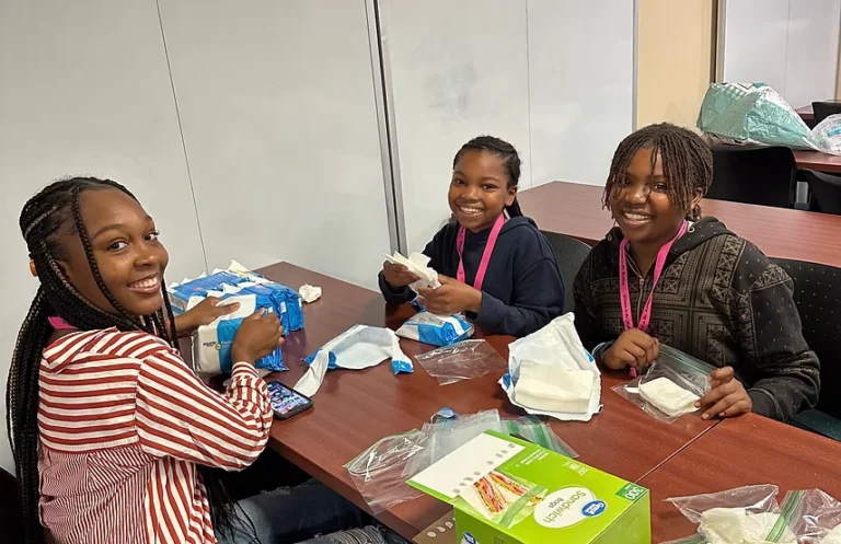 Three smiling young volunteers pack wipes and supplies into sandwich bags at a Packs of Love Foundation table.