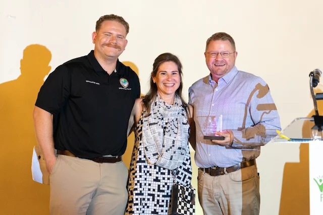 Two men and a woman smiling on a stage, one of the men holding a glass award.