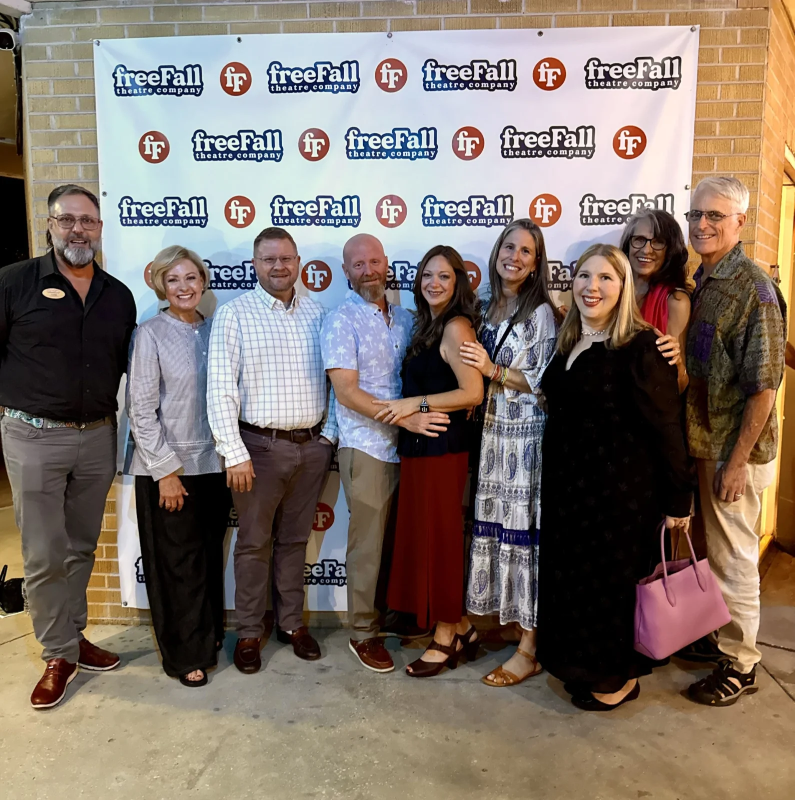 Nine adults smiling together in front of a freeFall Theatre Company step-and-repeat banner outside a brick building.