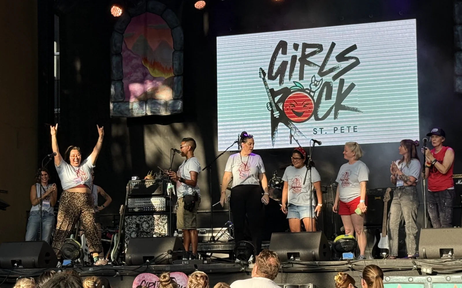 Volunteers in matching T-shirts on an outdoor stage at a Girls Rock St. Pete event, one with both arms raised, in front of a screen showing the Girls Rock St. Pete logo.