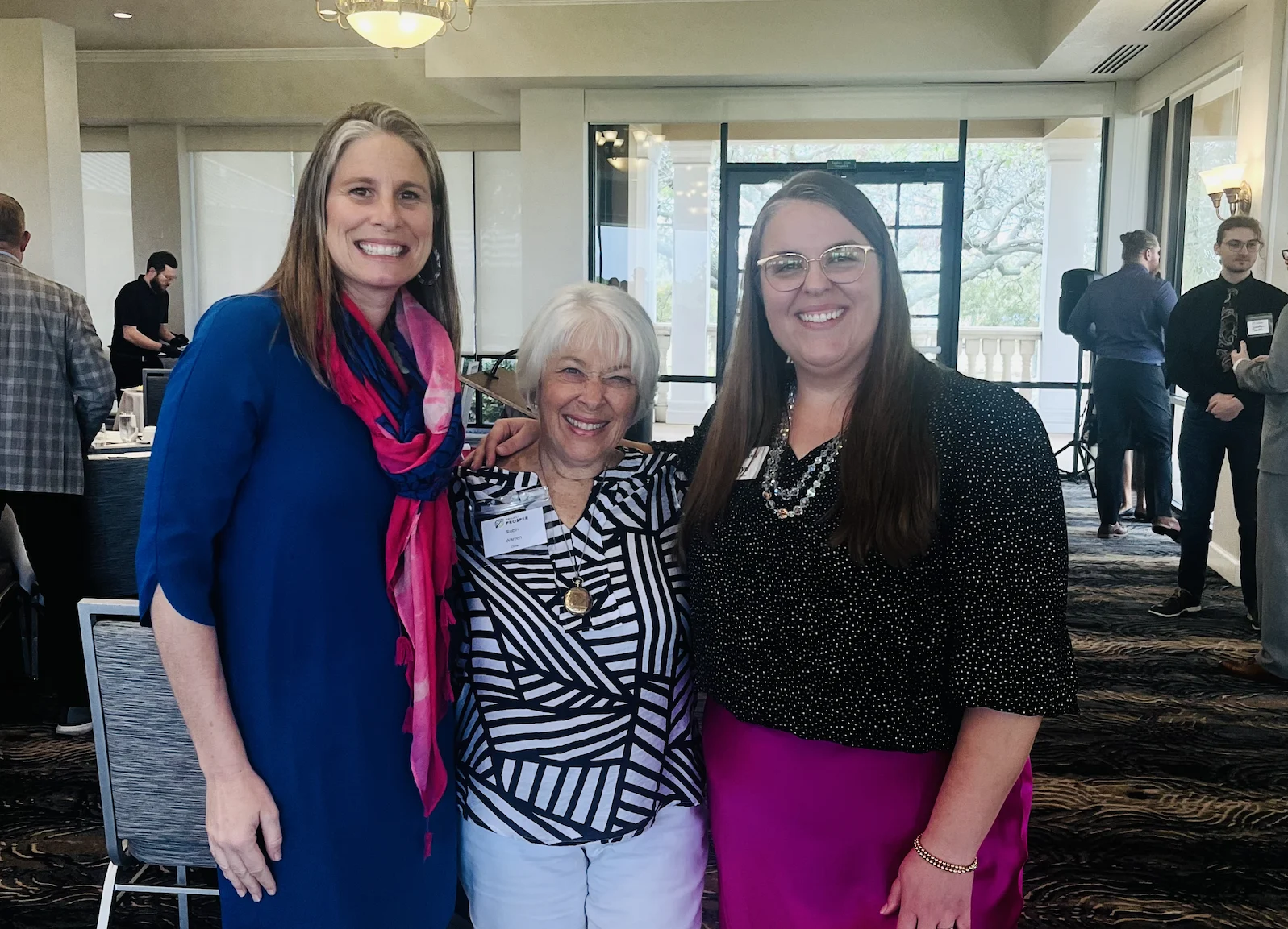 Three women smiling together at a luncheon in a hotel ballroom.
